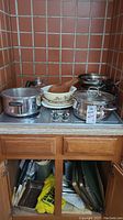 Photo of assorted pots and pans on stovetop and items below the kitchen cabinet including baking sheets and other cooking accessories.