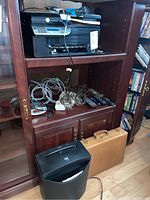 Wooden shelving unit with HP Officejet 4500 printer on top shelf, shredder on floor, and various cords and remotes on middle shelf.