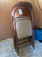 Four copper-coloured folding chairs stacked showing vinyl padded backs and seats with a beige tone, metal frames with wear.