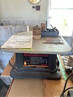 Oscillating spindle sander with sanding drums, accessories, and owner's manual on top mounted on plywood.