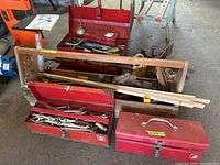 Photo of wooden tool carrier filled with various long hand tools and yardsticks, next to two red metal toolboxes on garage floor.