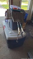 Photo showing stacked large resin tote boxes with hinged locking lids. A brownish work coat and pants are laid over the totes. Two ski poles with white handles are positioned beside the coat and pants.