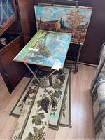 Four metal-framed TV trays each with scenic images and the grapevine patterned rugs beneath them on the floor
