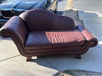 Full front side view of the brown leather antique chaise lounge on pavement, showing overall shape, leather upholstery, wooden studs, and legs.