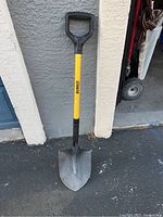 Full view of a DeWalt shovel standing against a wall showing the entire yellow handle, black grip, and pointed metal blade.