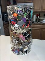 Large glass container densely filled with assorted jewelry pieces from front view, showing pearls, crystals, brooches, and colorful accessories.