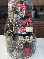 Photo showing side of glass pickle jar densely packed with mixed jewelry pieces inside including pearl necklaces, metal chains, and fabric-wrapped items.
