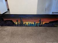 Full front view of the rectangular desert landscape painting showing the sunset, city skyline, and cacti silhouetted against the sky.