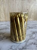 Front view of clear cylindrical container showing the gold taper Christmas tree candles inside, along with gold star decorations and brand text.