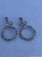 Pair of silver-tone metal hoop earrings decorated with clear rhinestones, featuring both round and baguette cuts around the circular frame, and screw back fasteners with an additional rhinestone near fastening.