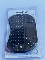 Front view of the iPazzPort mini wireless keyboard in original plastic packaging showing the QWERTY keyboard layout, touchpad, and directional buttons.