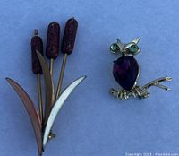Close-up of two brooches on a blue background: an owl brooch with green eyes and purple body stone, and a cattail plant brooch with three reddish-brown cattails and metal leaves.