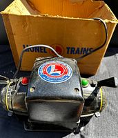Front and top view of the black metal Lionel Type-ZW transformer with logo sticker, control knobs, and attached power cable, set in front of the original box.