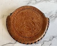 Front view of round brown leather coin purse showing embossed Mexican design with figure, sun, coins, and "Mexico" text.