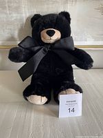 Front view of the black teddy bear sitting upright with a large black satin bow tied around the neck. Fur appears soft and plush with light brown feet visible.