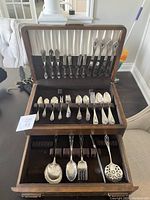 Top view of the open antique wooden box showing multiple rows of silver flatware including forks, spoons, and knives arranged in velvet slots.