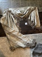Pair of asymmetrical beige/grey valance drapes laid out on floor; floral pattern on one side with fringed edges and pinstripe reverse