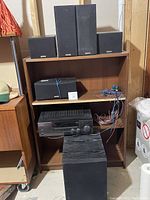 Image showing all speakers and the tuner/amplifier on shelving. Includes one large speaker, three tall speakers, two small speakers and tuner/amplifier; black enclosures with fabric covers.