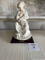 Front view of the white porcelain sculpture of a mother breastfeeding an infant on wooden base, showing detailed features and clothing folds.