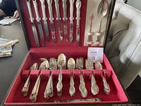 Open wooden box showing two rows of knives, forks, spoons, and serving utensils arranged in a red velvet interior.