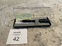Black digital recording pen in an open plastic case with clear lid, resting on protective foam inside the case. A handwritten note labeled 'Radio Shack Digital Recording Pen' is included inside the case alongside instructions.