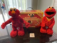 Both red plush Tickle Me Elmo robotic toys side by side with the original Extra Special Edition box behind them on a table in daylight.