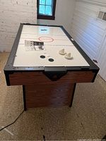 Full view of air hockey table showing white playing surface with red and blue markings, two white paddles, three pucks, and owner's manual on table. Wood patterned sides and black rails visible.