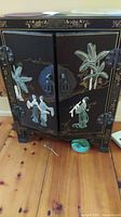 Front view of black Asian style cabinet with two doors depicting raised designs of people and plants. Cabinet has a circular metal lock and decorative metal hinges.
