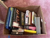 Cardboard box filled with assorted books, including yearbooks and fiction titles visible from top-down view showing variety of book sizes and covers