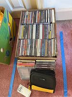 Top-down view of a cardboard box filled with multiple music CD jewel cases and some CDs stacked outside the box near a black and yellow CD wallet pouch.