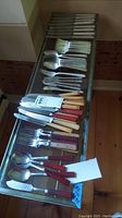 Overview of assorted knives, forks, and spoons with assorted handle materials laid out on table.