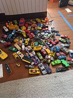 Photo of a large assortment of diecast cars, plastic trucks, and military vehicles scattered on floor showing variety of colors and vehicle types.