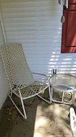 Patio rocker and glass top table placed on a concrete porch against white siding, shows dirt, leaves, and sunlit patches on the surfaces.
