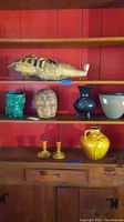 Shelf view of lot showing Egyptian mummy replica, Roseville pottery vases, green planter, brown clay head, and yellow candle holders.