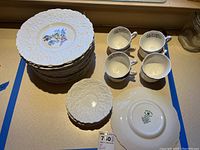 Stack of 9.25 inch embossed white plates with floral center designs, 4 cups with matching floral patterns, and a plate showing the Royal Cauldon backstamp.
