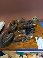 Classic vintage metal irons with wooden handles, electric iron, and brass lamp parts on wooden table.