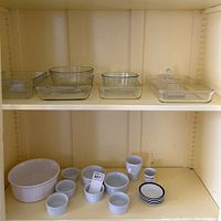 Shelf with multiple clear Pyrex glass baking dishes including rectangular and square shapes, plus a clear crystal butter dish; below, assorted white ceramic bowls and some small cups and plates.