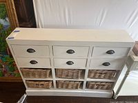Front view of the white wooden cabinet showing its six drawers on upper half with black cup handles and six wicker baskets below.