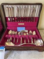 Full view of open display case showing entire silverware set with knives, forks, spoons, and serving pieces arranged in red velvet and satin-lined compartments.