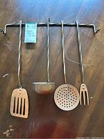 Copper cooking utensils including slotted spatula, ladle, skimmer, and fork hanging on twisted metal rack laid on wooden table.