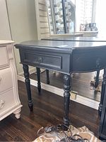 Side angle view showing a black vintage wood console table with a single drawer, turned legs, and some distressing on the finish.