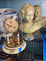 Photo showing German-made table clock under glass dome with floral decoration on clock face and base, placed next to two resin busts