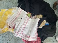 Pile of assorted linens including folded white tea towels with red stitching, yellow checkered towels, and black napkins with a black rolling suitcase visible