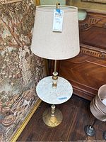Front and side view of marble table lamp showing white marble disc on stand, brass base, beige fabric lampshade, and overall design