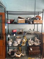 Full front view of the Metro metal shelving rack loaded with various kitchenware items including ceramic pitchers, bowls, glass dome covers, and brown vintage style suitcases on the bottom shelf.