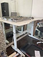 Right-off front angle view of white modern adjustable desk showing desk top with two decorative trays and part of surrounding furnishings.