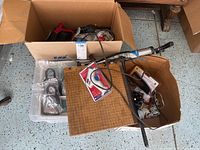Wide shot showing cardboard boxes containing assorted items including a wooden paper cutter, bike locks, drill, and other miscellaneous goods