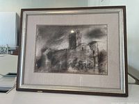 Framed artwork showing a dark castle scene with prominent tower, surrounded by trees, displayed on a table indoors.