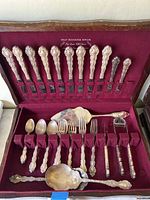 Partial silverware set with 10 dinner knives and several serving utensils resting in a velvet-lined wooden box with '1847 Rogers Bros.' printed inside the lid.