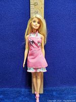 Barbie doll standing against blue background with ruler for size reference. The doll is wearing a pink apron over a patterned sleeveless dress and pink high-heeled shoes.